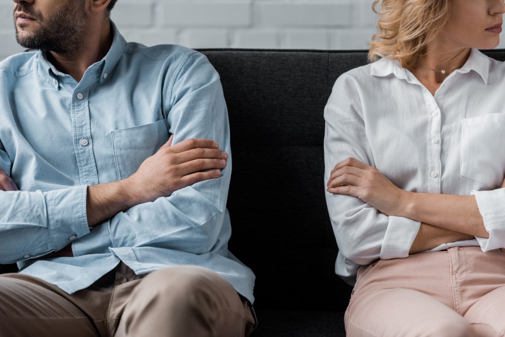 Two adults sitting back-to-back on a dark couch with arms crossed; man in light blue shirt left, woman in white blouse right.