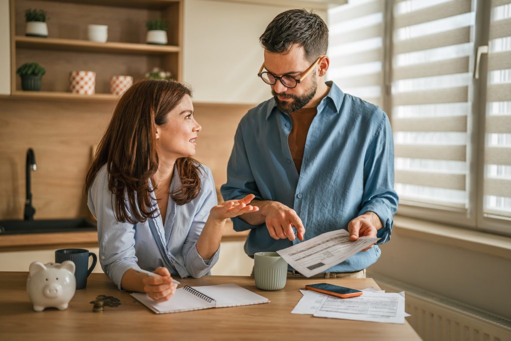 Two people in a kitchen discuss documents on a table, with notebooks, a receipt sheet, and mugs nearby.